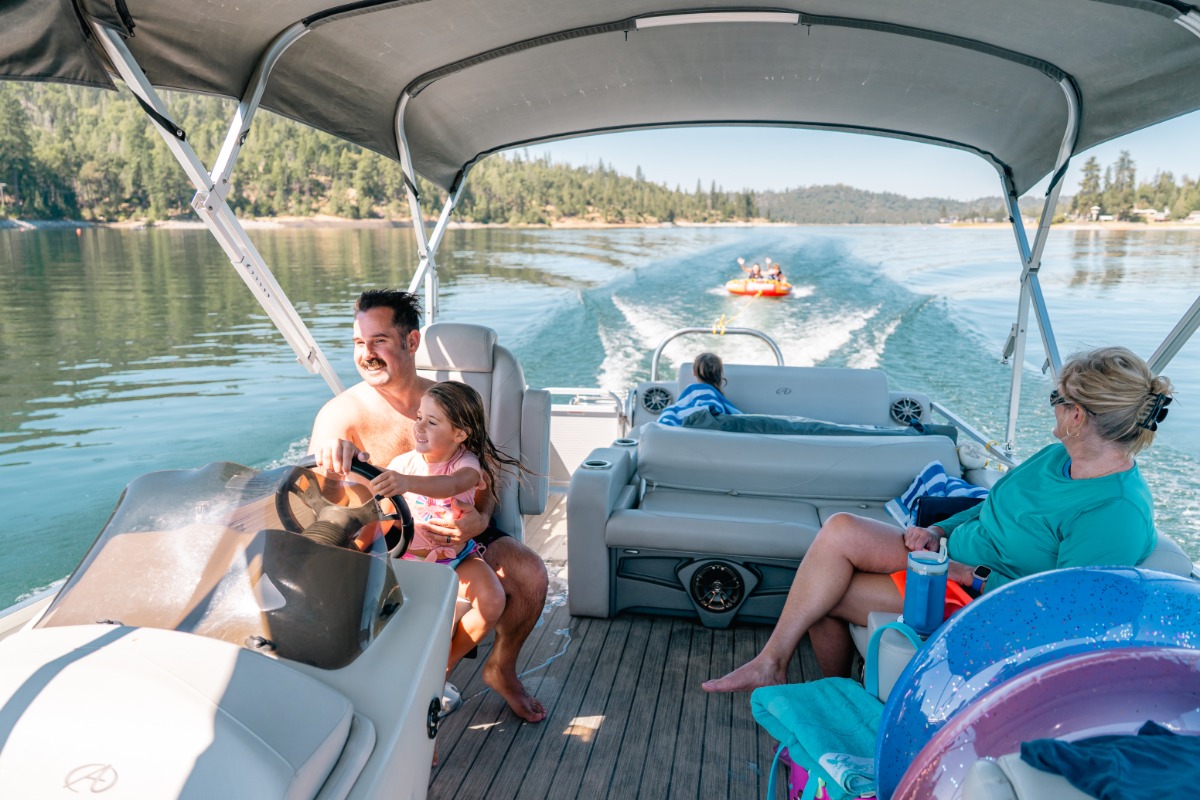 Man drives pontoon boat with family, mother this intertubing behind the boat.
