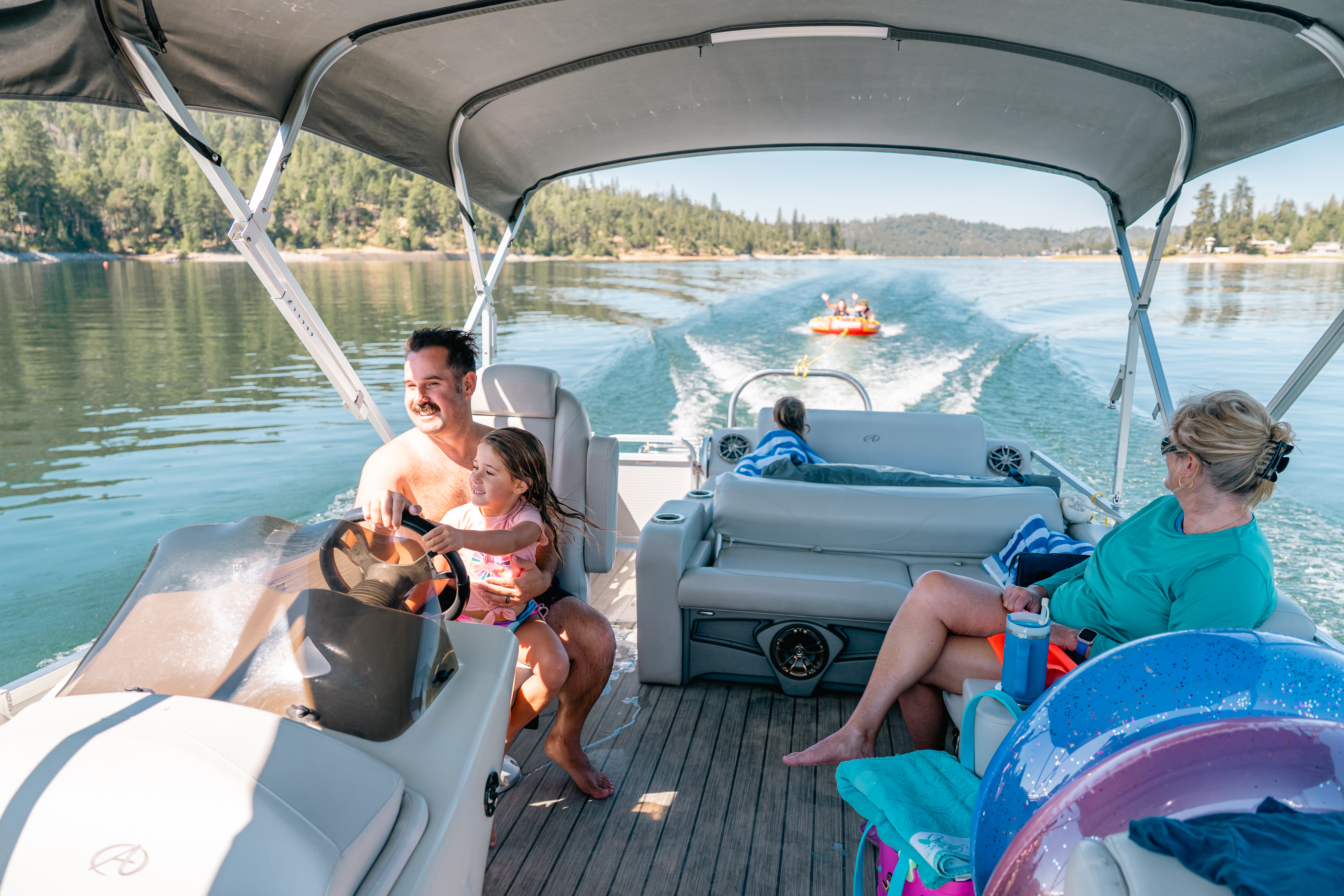 Man drives pontoon boat with family, mother watches kids intertubing behind the boat. 